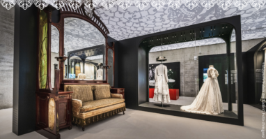 Fabrik der Fäden Das Bild zeigt einen Ausstellungsraum mit einem Sofa im Vintage-Stil und zwei Schaufensterpuppen, die aufwendige, historische Kleidungsstücke tragen. Im Hintergrund sind dekorative Muster an der Decke und den Wänden zu sehen, die eine elegante Atmosphäre schaffen.