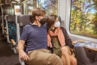 Ein Pärchen mit Masken sitzt im trilex nebeneinander und schaut aus dem Fenster.