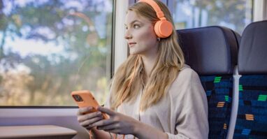 Eine junge Frau sitzt mit Handy und Kopfhörern im trilex und hört Musik, während sie aus dem Fenster schaut.